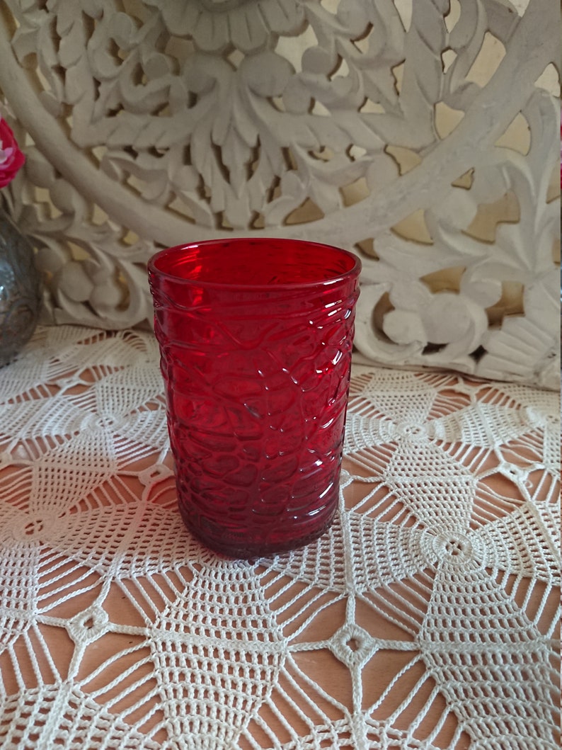 May include: A red glass tumbler with a textured pattern. The tumbler is sitting on a white crocheted tablecloth.