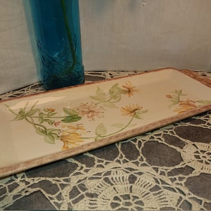 Può includere: Un vassoio rettangolare in ceramica con un design floreale. Il vassoio ha uno sfondo beige chiaro con un bordo rosa e un motivo floreale di fiori gialli e rosa con foglie verdi.