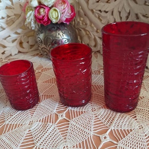 May include: Three red glass tumblers of varying sizes with a textured, woven pattern. The tumblers are arranged in a row from smallest to largest on a white crocheted tablecloth.