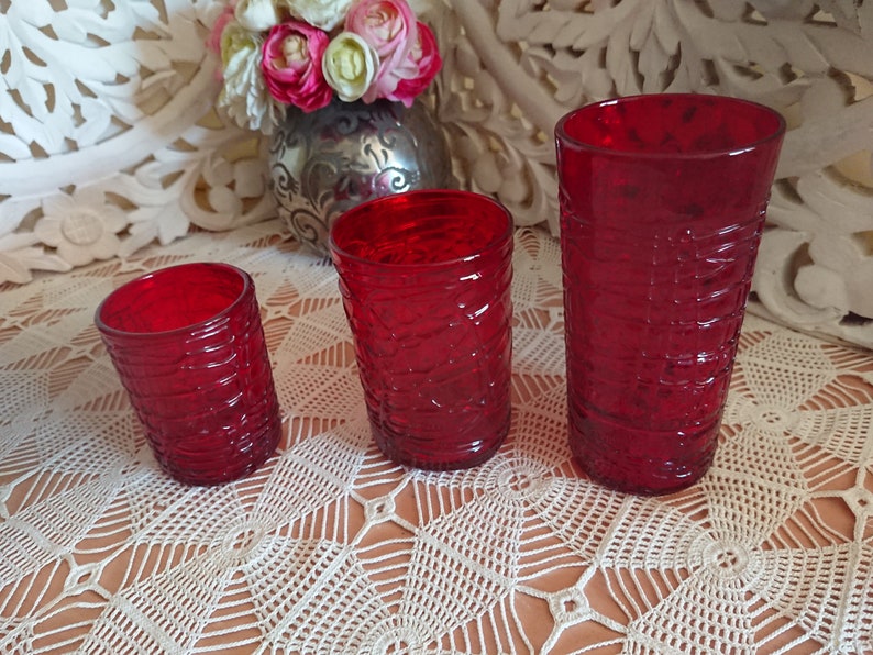 May include: Three red glass tumblers of varying sizes with a textured, woven pattern. The tumblers are arranged in a row from smallest to largest on a white crocheted tablecloth.