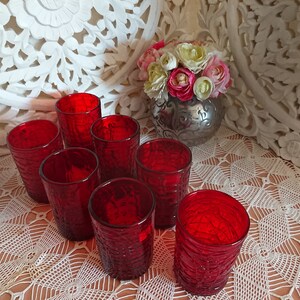 May include: Eight red glass tumblers with a textured surface. The glasses are arranged in a circle on a white crocheted tablecloth.