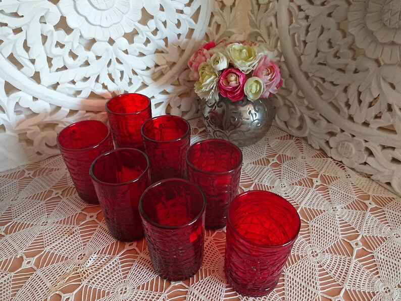 May include: Eight red glass tumblers with a textured surface. The glasses are arranged in a circle on a white crocheted tablecloth.