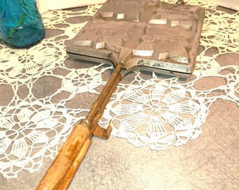 Op de afbeelding: Een vintage wafeltang met een houten handvat. De tang heeft vier vierkante wafelvormen en is gemaakt van metaal. De tang ligt op een wit gehaakt doily.
