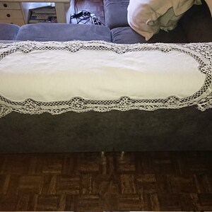 May include: White crocheted table runner with a delicate lace pattern. The runner is laid out on a gray couch.