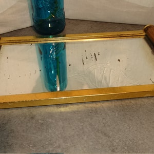 Op de afbeelding: Rechthoekige spiegel met een messing frame en houten handvatten. De spiegel heeft een goudkleurig frame en bruine houten handvatten met messing accenten. Een blauwe glazen fles wordt weerspiegeld in de spiegel.