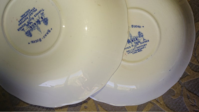Op de afbeelding: Twee witte keramische schoteltjes met blauwe en witte ontwerpen. De schoteltjes zijn afkomstig van de Palissy Pottery in Staffordshire, Engeland. De tekst op de schoteltjes luidt "Bray Bucks" en "Bucks: Thames River Scenes Palissy Pottery Staffordshire England."
