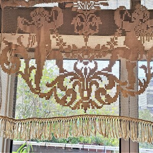 May include: A white crocheted valance with a brown floral design and a fringe trim. The valance is hanging in a window.
