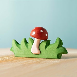 A vibrant red and white spotted wooden mushroom toy emerging from a green wooden grass base, set against a soft blue background.
