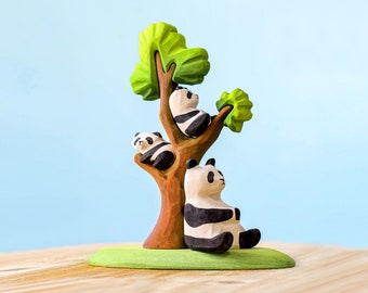 Peut inclure: Un arbre en bois jouet avec trois pandas en bois sculptés. Les pandas sont noirs et blancs et sont assis sur les branches de l'arbre. L'arbre est sur une base verte.