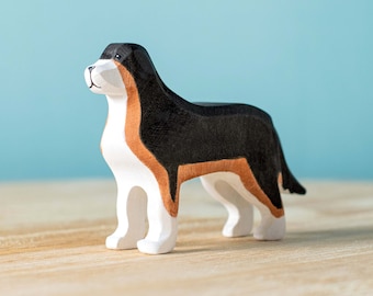 May include: A wooden toy dog figurine with black, white, and brown markings. The dog is standing on a light wood surface.
