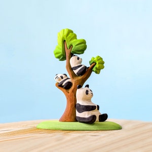 Op de afbeelding: Een houten speelgoedboom met drie gegraveerde pandaberen. De pandaberen zijn zwart en wit en zitten op de takken van de boom. De boom staat op een groene basis.