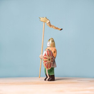 A wooden knight figurine with a hand-painted shield and a tall lance topped with a whimsical fox head banner, standing on a wooden surface against a soft blue background.