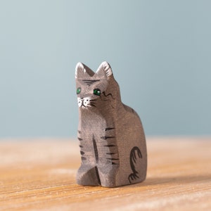 Handcrafted grey wooden cat figure with green eyes and black stripes standing on a wooden surface, Waldorf inspired toy for animal education.