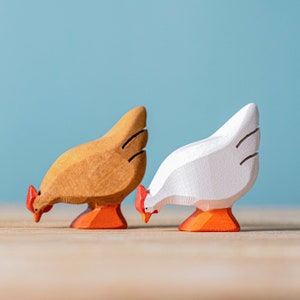 A wooden rooster and hen toy side by side, both with red combs and orange feet, the rooster with a detailed brown body and the hen in white, against a soft blue background.
