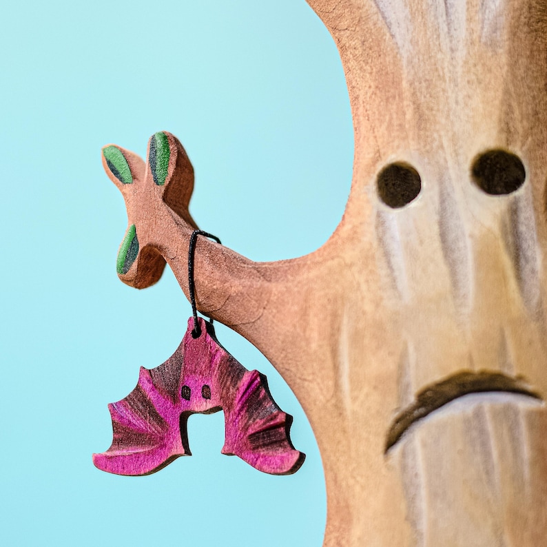 Close-up view of a wooden bat figurine hanging from the branch of a handcrafted tree toy with textured bark and somber expression.