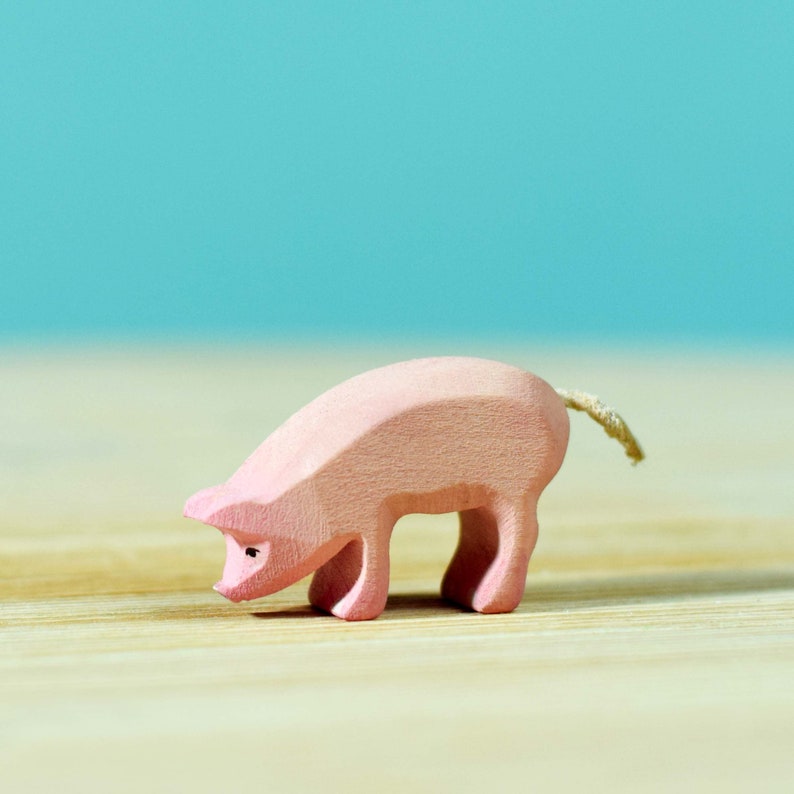 A small wooden piglet figure, delicately painted in light pink, standing on a wooden surface with a soft-focus turquoise background.