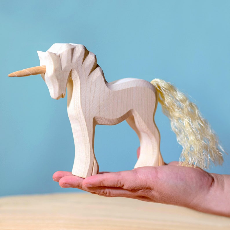 Handcrafted wooden unicorn held in a person's hand, showcasing the toy's size and detailed mane and horn against a pale blue background.