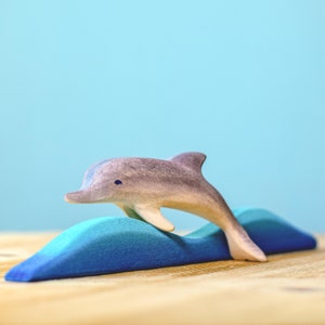 A singular wooden dolphin arches gracefully over a blue felt wave, showcasing the simplicity and charm of BumbuToys' handcrafted sea creatures against a calm blue sky.