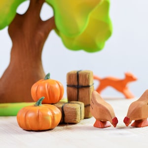 A whimsical wooden toy scene with two hens pecking near wooden pumpkins and a fence, under a large green tree on a light wood table.