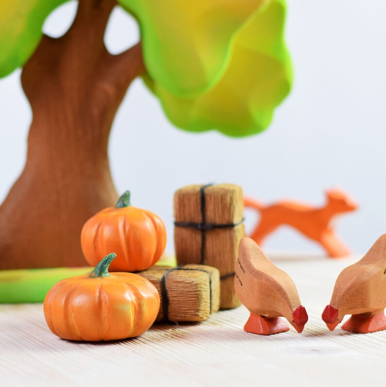 A whimsical wooden toy scene with two hens pecking near wooden pumpkins and a fence, under a large green tree on a light wood table.