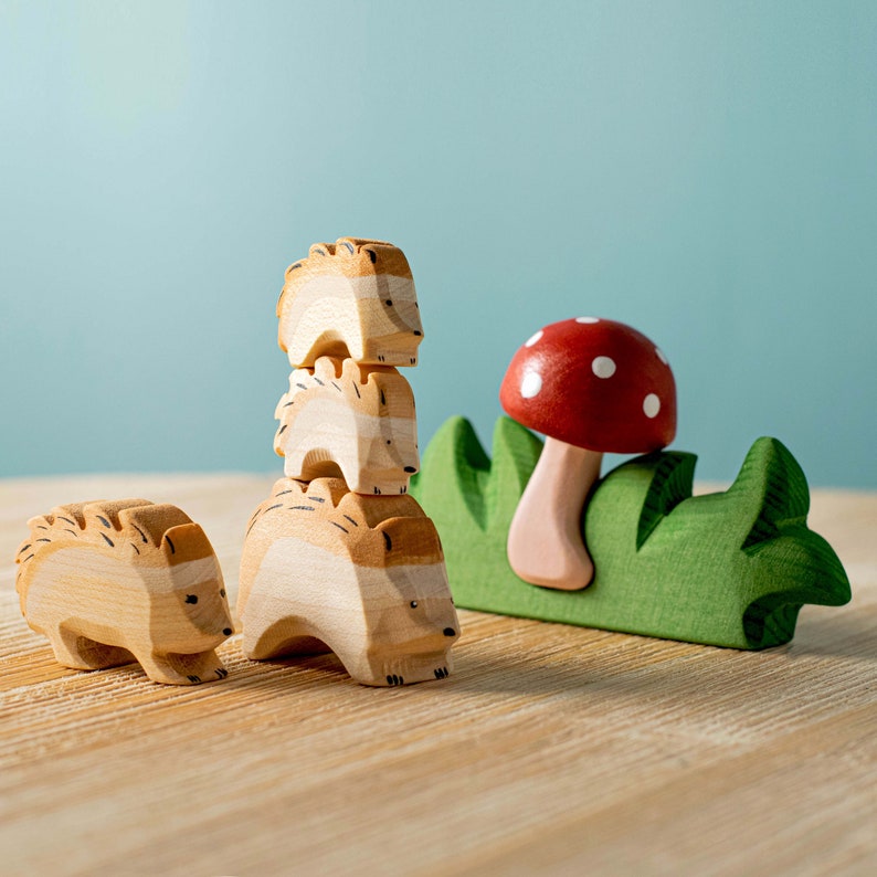 Three wooden hedgehog toys in different sizes lined up in front of a red mushroom with green grass, on a wooden table.