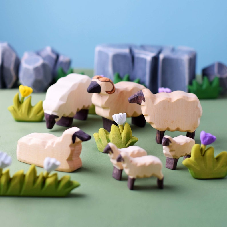 Handcrafted wooden sheep toys arranged on a green surface with colorful flowers and grey rock formations in the background.