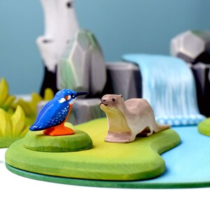Wooden kingfisher toy beside a wooden otter and river, part of a BumbuToys playset that encourages storytelling and exploration of river ecosystems.