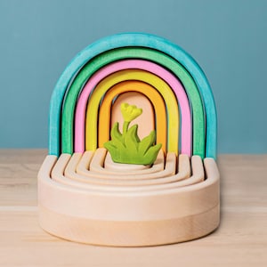 May include: A rainbow-colored wooden arch toy with a small green and yellow flower in the center. The arch is made of seven interlocking pieces, each a different color of the rainbow.