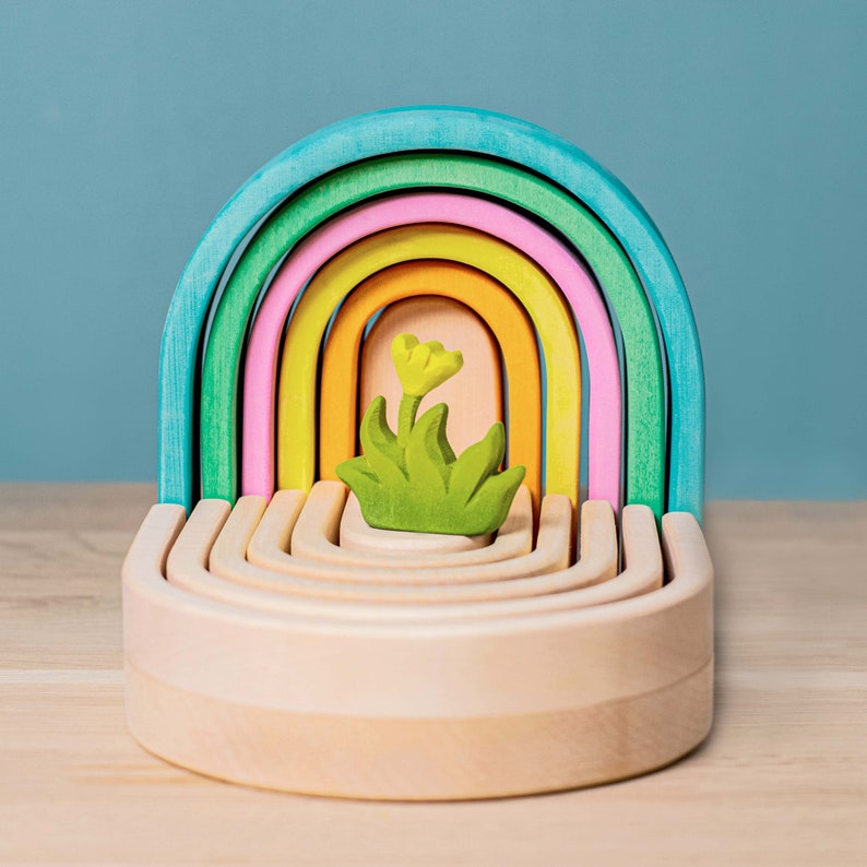 May include: A rainbow-colored wooden arch toy with a small green and yellow flower in the center. The arch is made of seven interlocking pieces, each a different color of the rainbow.