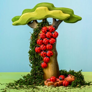 A whimsical arrangement of red wooden ladybugs on a tree branch with a large green leaf overhead, set against a soft blue sky.