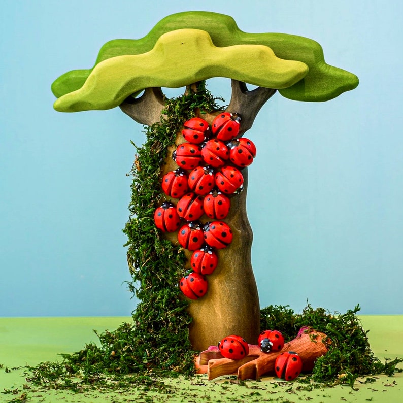 A whimsical arrangement of red wooden ladybugs on a tree branch with a large green leaf overhead, set against a soft blue sky.