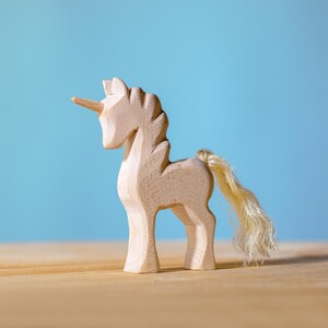 Front-facing view of a wooden unicorn with a spiral horn and wavy tail, placed on a light wooden surface with a sky-blue background.