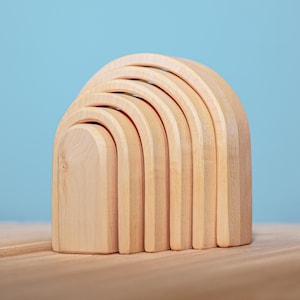 May include: A set of seven natural wooden rainbow arches, stacked in a semi-circle shape. The arches are made of unfinished wood and have a smooth, rounded surface.