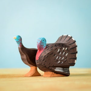 Op de afbeelding: Twee houten kalkoenfiguren, een met een blauwe kop en de andere met een rode kop, staan op een houten oppervlak. De kalkoen met de blauwe kop kijkt naar links en de kalkoen met de rode kop kijkt naar rechts.