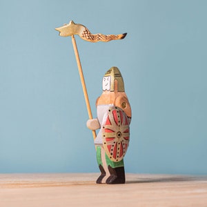 Handcrafted toy knight with detailed painting, holding a lance with a fox banner, shown from a different angle with a blue backdrop, on a wooden surface.