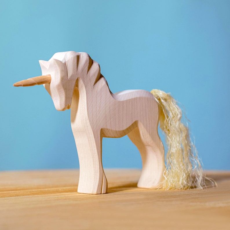 Wooden unicorn figurine with spiral horn and fluffy tail, standing on a wooden surface against a pale blue background.