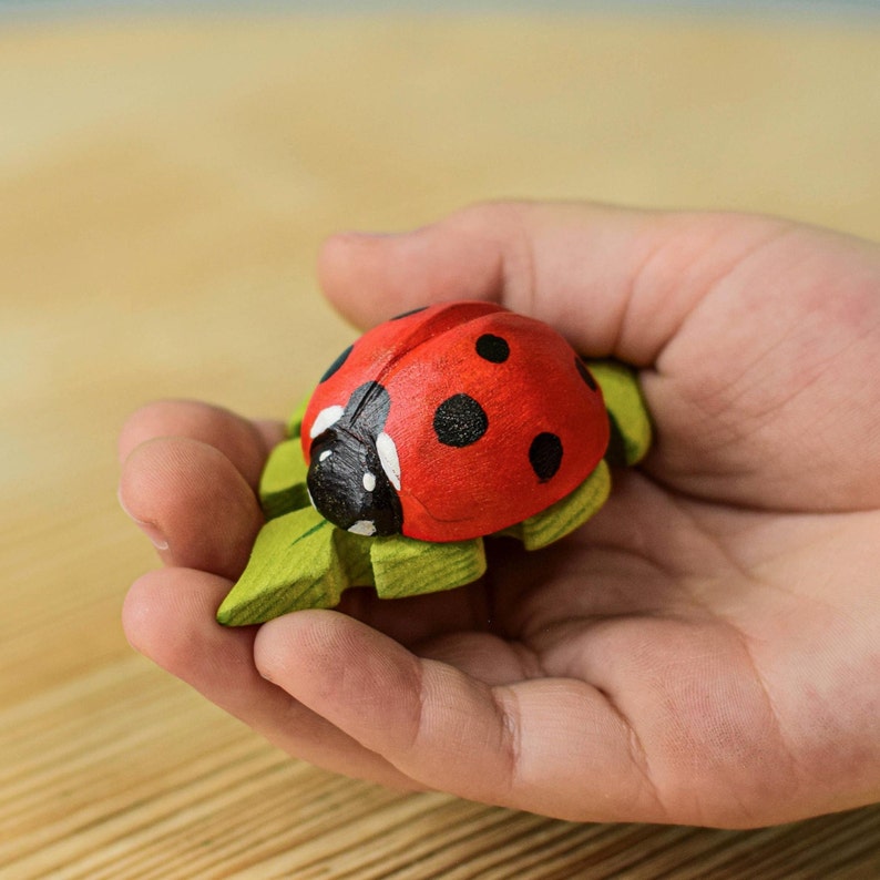 A child's hand gently holding a hand-painted wooden ladybug toy with vivid red wings and black spots on a leaf-green base.