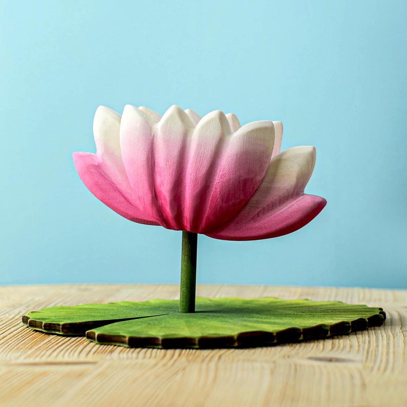 A wooden lotus flower sculpture with pink and white petals, standing on a green lily pad against a soothing blue background.