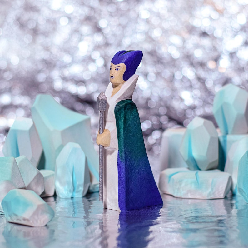 A wooden figurine with purple hair and a green and white outfit holding a staff, standing among ice-like wooden blocks with a glittery background.