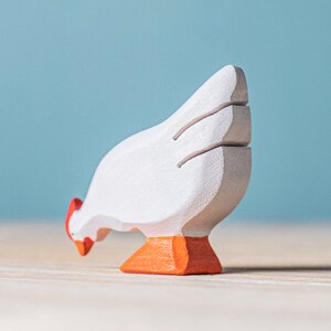 Side profile of a white wooden hen toy with red detailing on the comb, positioned on a wooden surface with a plain blue background.