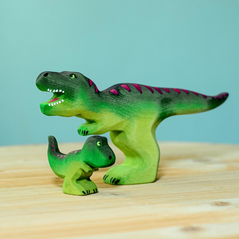 Two wooden T-Rex toys painted in green with magenta back stripes, positioned as if interacting, on a wooden table against a teal background.