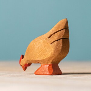 Close-up of a wooden brown rooster toy with intricate black tail feather details, bent down pecking, set against a pale blue wall.