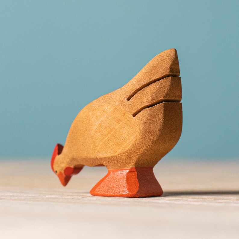 Close-up of a wooden brown rooster toy with intricate black tail feather details, bent down pecking, set against a pale blue wall.