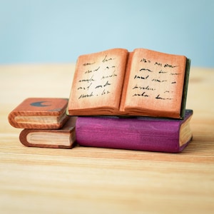 May include: A stack of three miniature books with brown covers. The top book is open and has handwritten text on the pages. The bottom book has a purple cover.