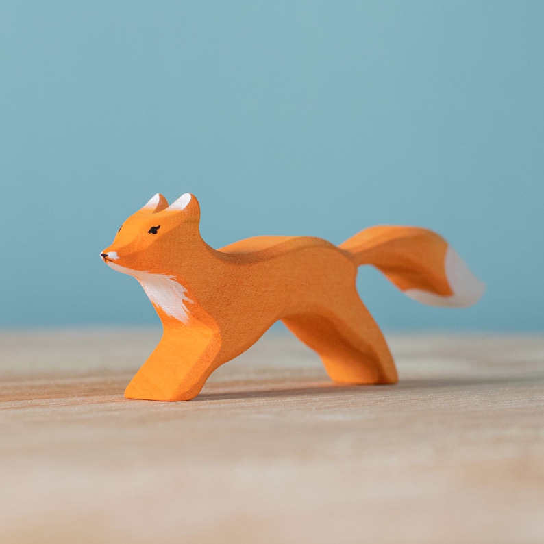 Wooden fox toy in a running pose with white accents on the body, presented on a wooden surface with a light blue background.