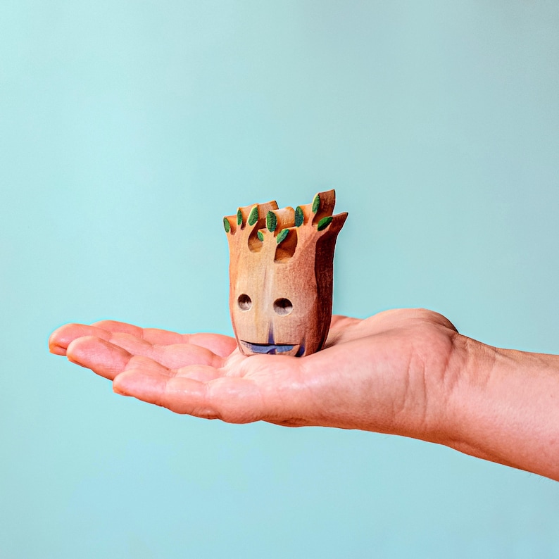 A person's hand gently holding a small, carved wooden tree stump with green leaves, highlighting the craftsmanship and scale of the Halloween toy.