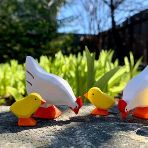 Three-dimensional wooden toy figures of a hen and chicks in natural outdoor lighting, with vibrant green foliage in the background.