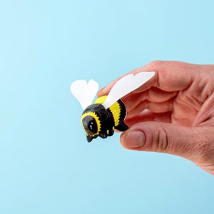 May include: A hand is holding a small, black and yellow bee figurine with white paper wings. The bee has a cute, cartoon-like face.