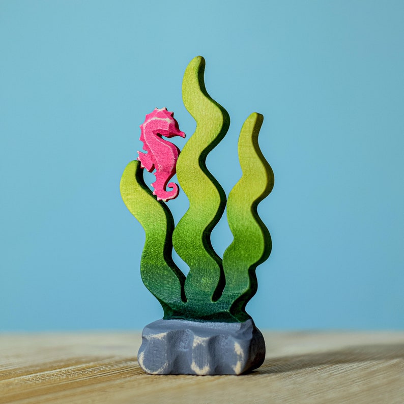Pink wooden seahorse figurine nestled in a vibrant green sea plant, the figure's color strikingly contrasted with the green shades.