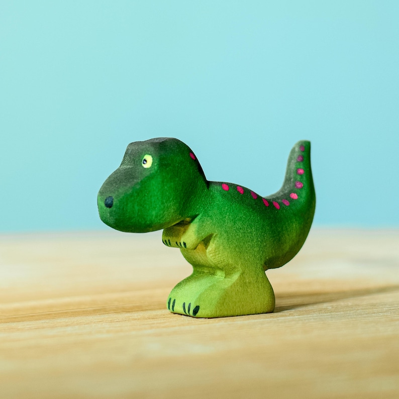 Small green wooden T-Rex toy with magenta spots along its back, standing solo on a wooden surface with a soft-focus teal background.